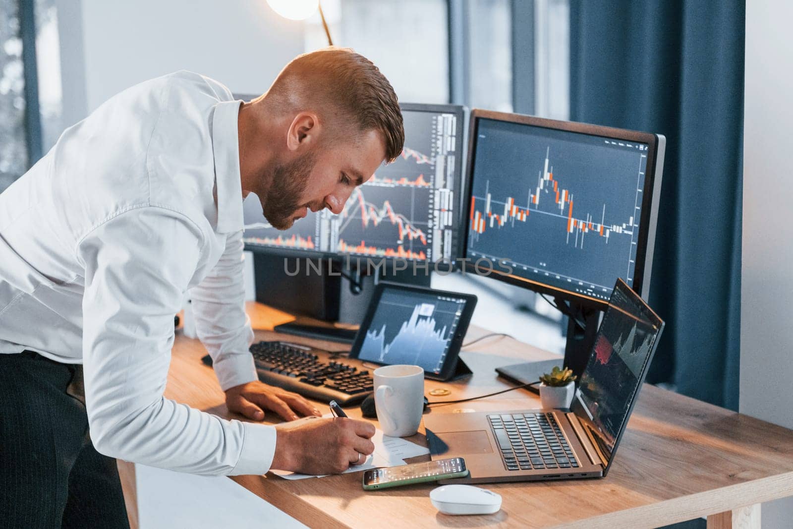 Near the table. Young businessman in formal clothes is in office with multiple screens.