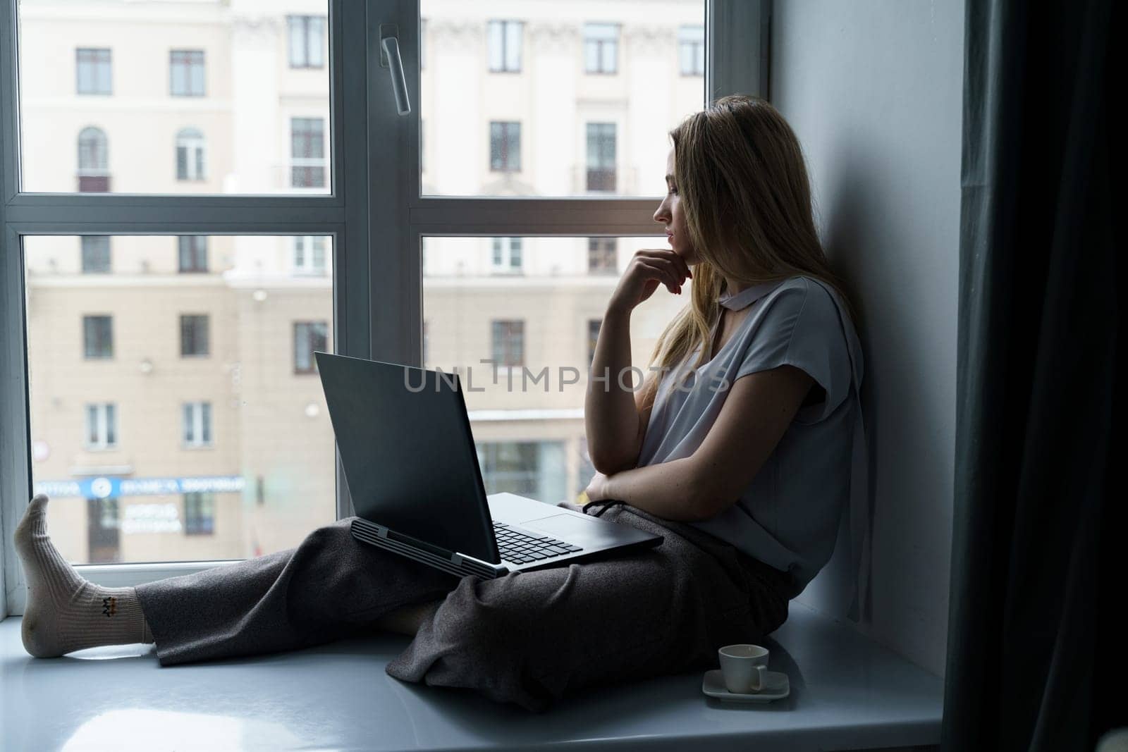 A young woman sits on a windowsill, works at a laptop, looks out the window. by Sd28DimoN_1976