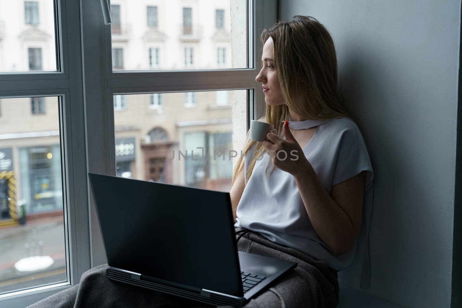 A young woman sits on a windowsill, drinks coffee, works at a laptop, looks out the window. Business and education concept. Close-up.