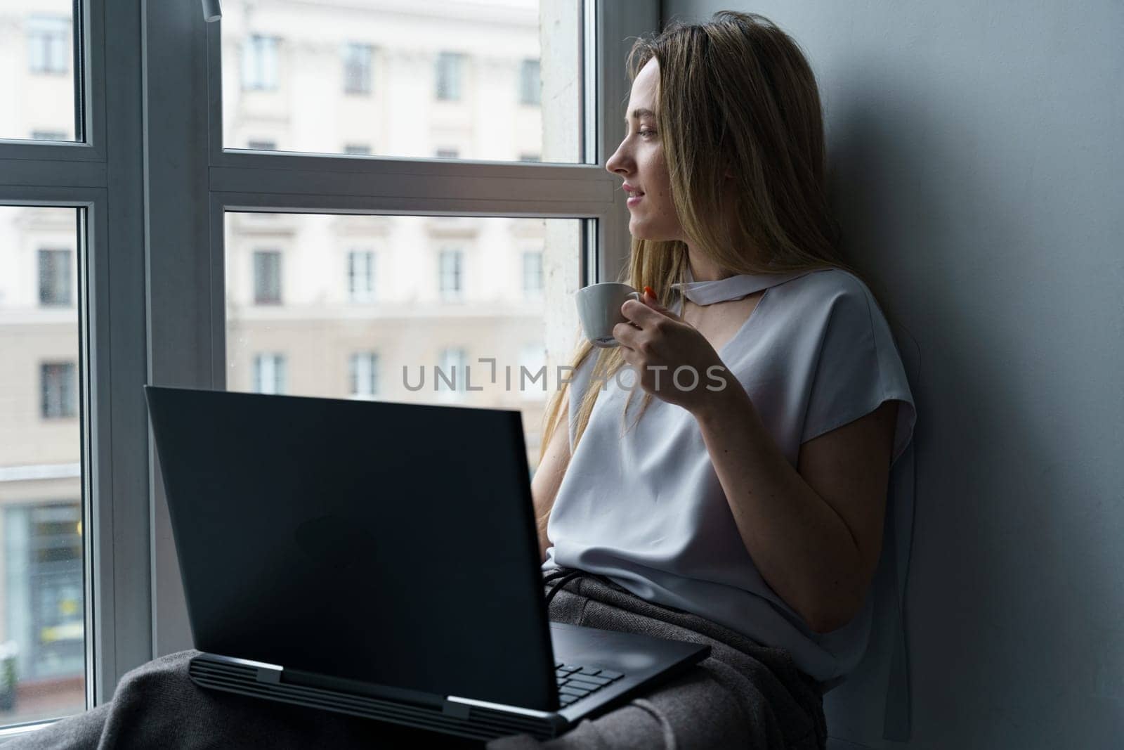 A young woman sits on a windowsill, drinks coffee, works at a laptop, looks out the window. by Sd28DimoN_1976