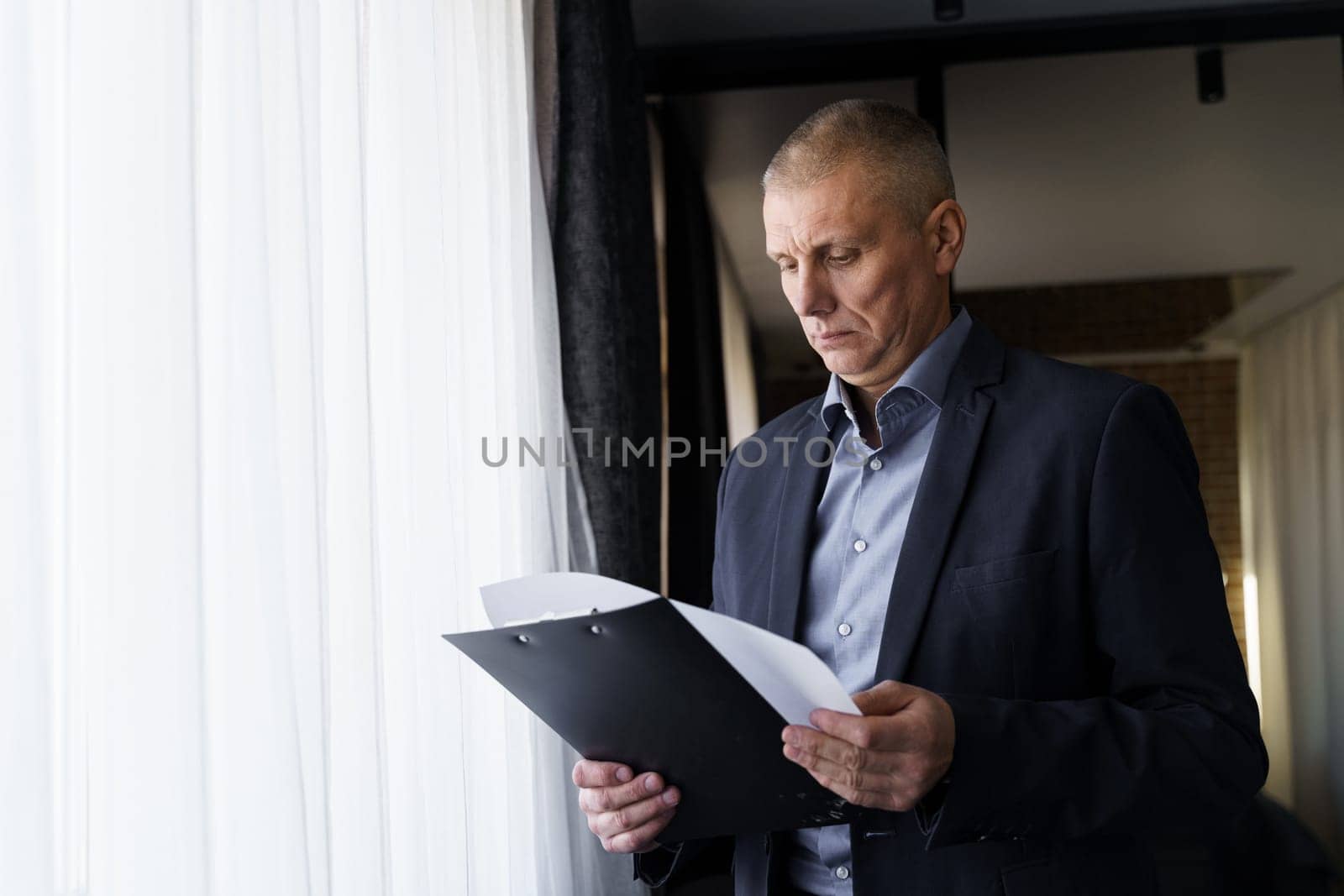 A middle -aged businessman stands near the window in the office and studies documents.
