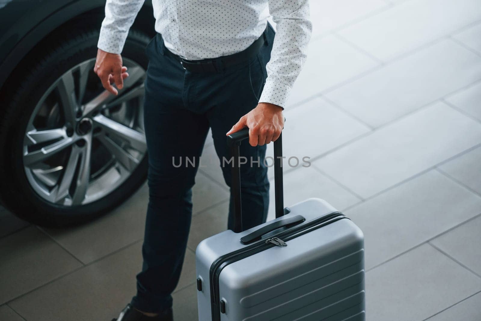 Partical view. Man in formal clothes is with luggage indoors near the car.