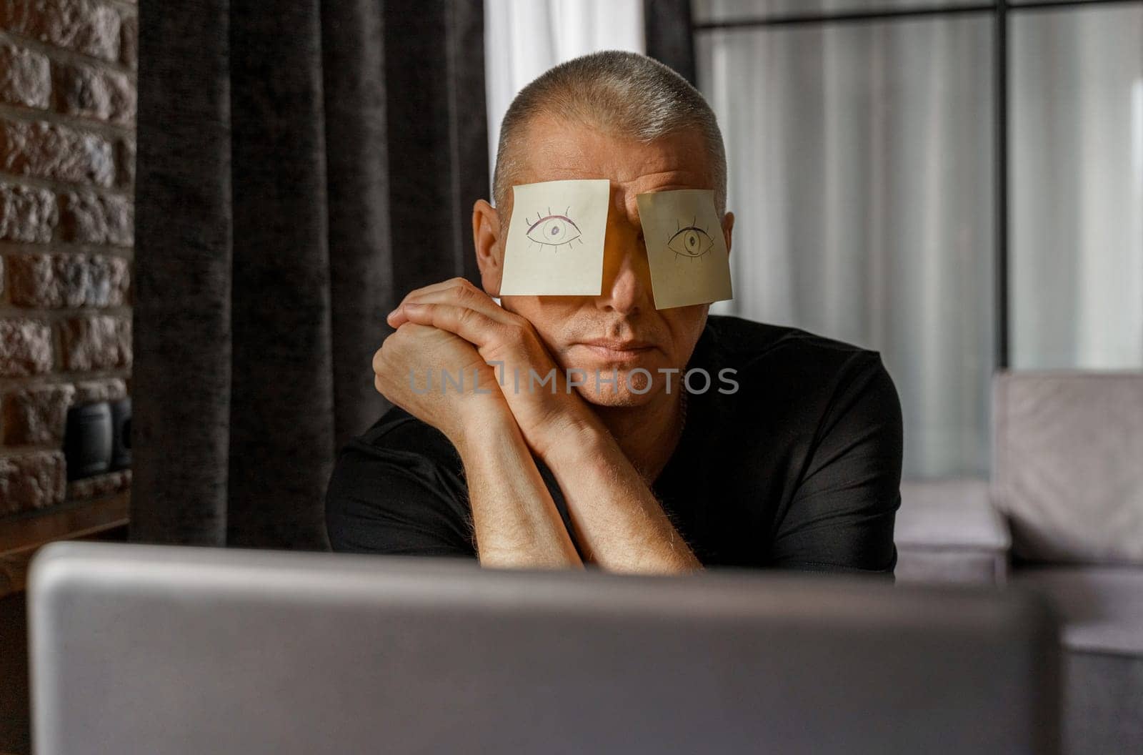 A tired businessman with stickers on his face is sitting at a table in the office, sleeping, eyes are drawn on the stickers.
