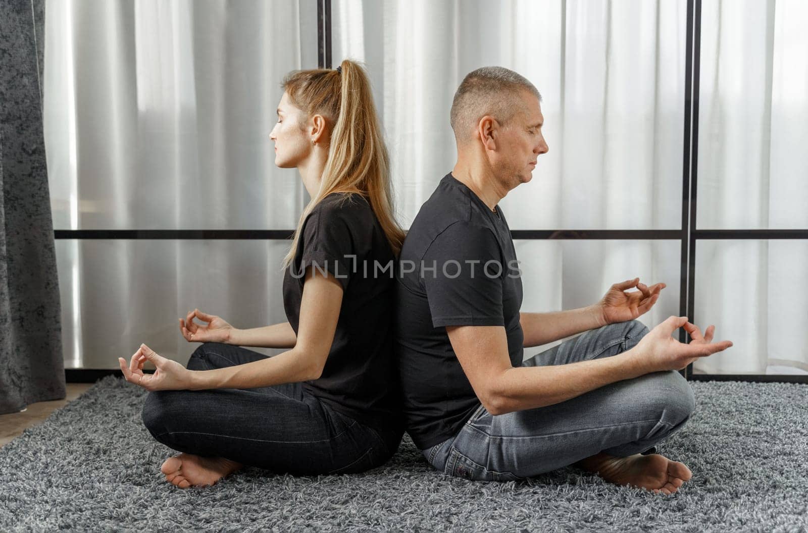 A man and a woman sit back to back, practice yoga and meditate for relaxation and balance of life in a room at home. by Sd28DimoN_1976