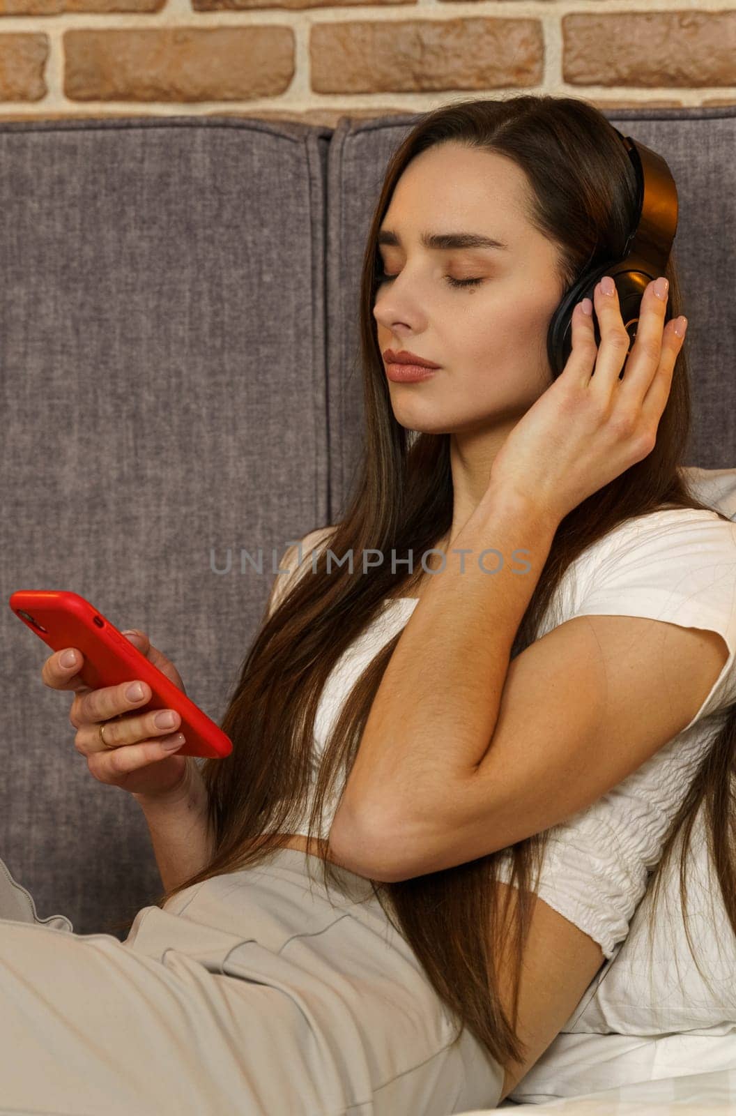 Happy young woman in headphones sits on the sofa and listens to music from a mobile phone, chooses tracks. Side view. Vertical frame.