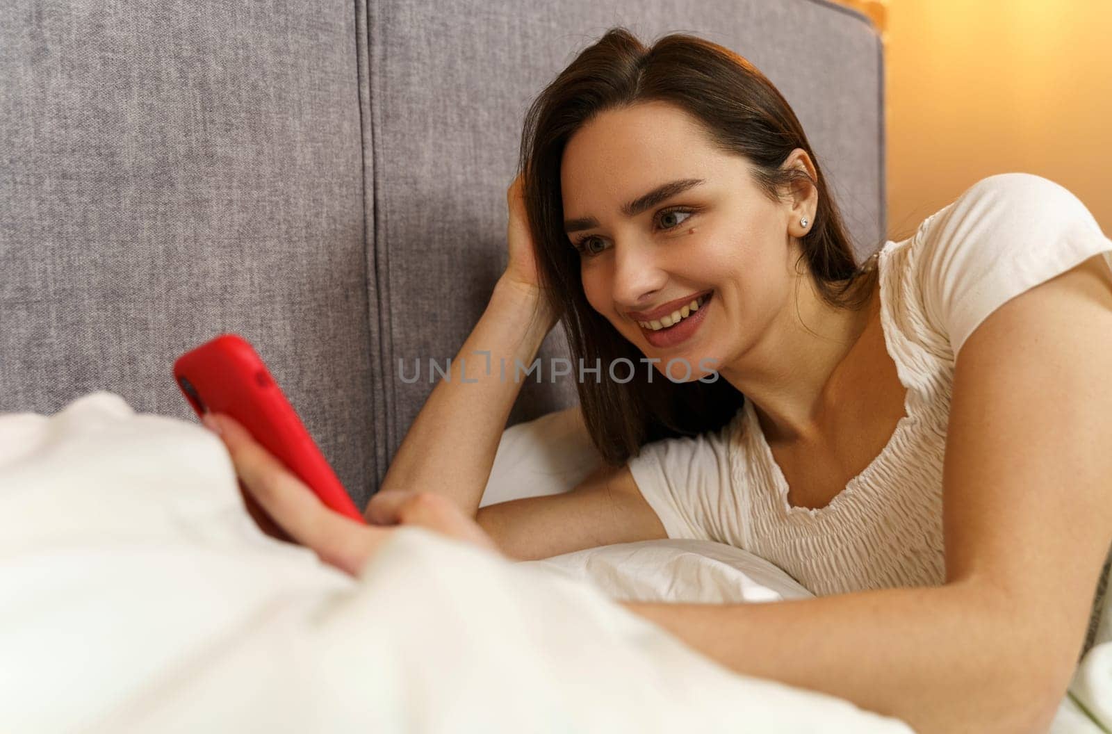 A beautiful young girl lies in bed and enjoys communication, holds a smartphone in her hands.