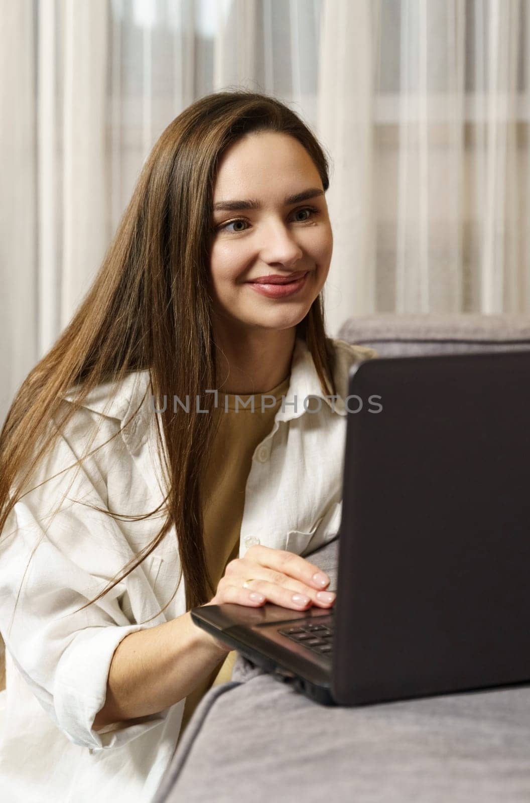 A beautiful young female freelancer is working on a laptop which is standing on the sofa, she is sitting on the floor herself.