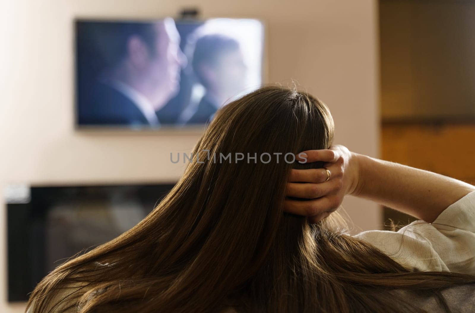 The girl watches TV, sits in the living room at home in the evening, switches TV channels. View over the shoulder, the image is blurry.