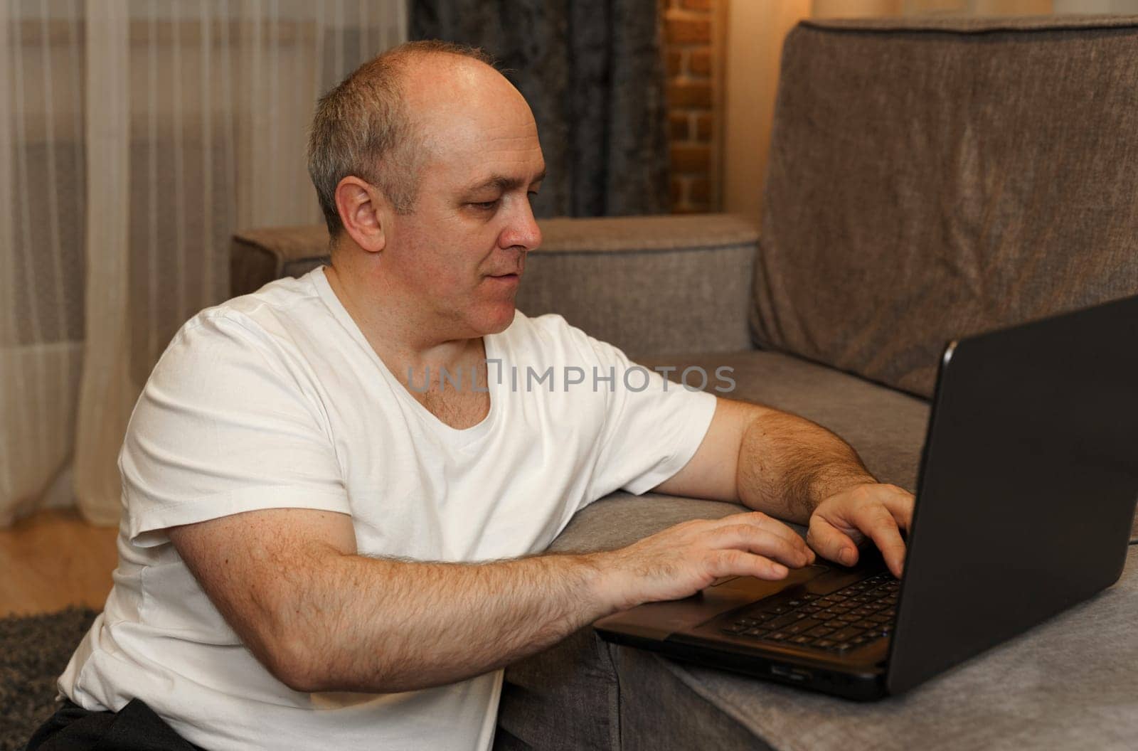 A mature man sits on the floor and works on a laptop that is on the sofa.