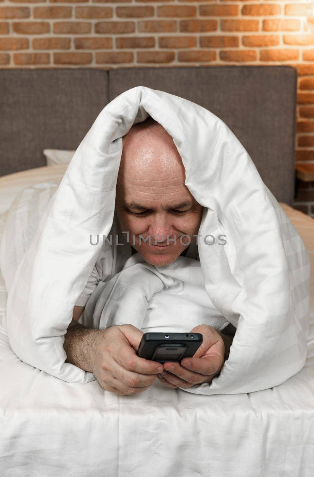 An adult man lies covered with a blanket and browses social networks, videos in a smartphone by Sd28DimoN_1976
