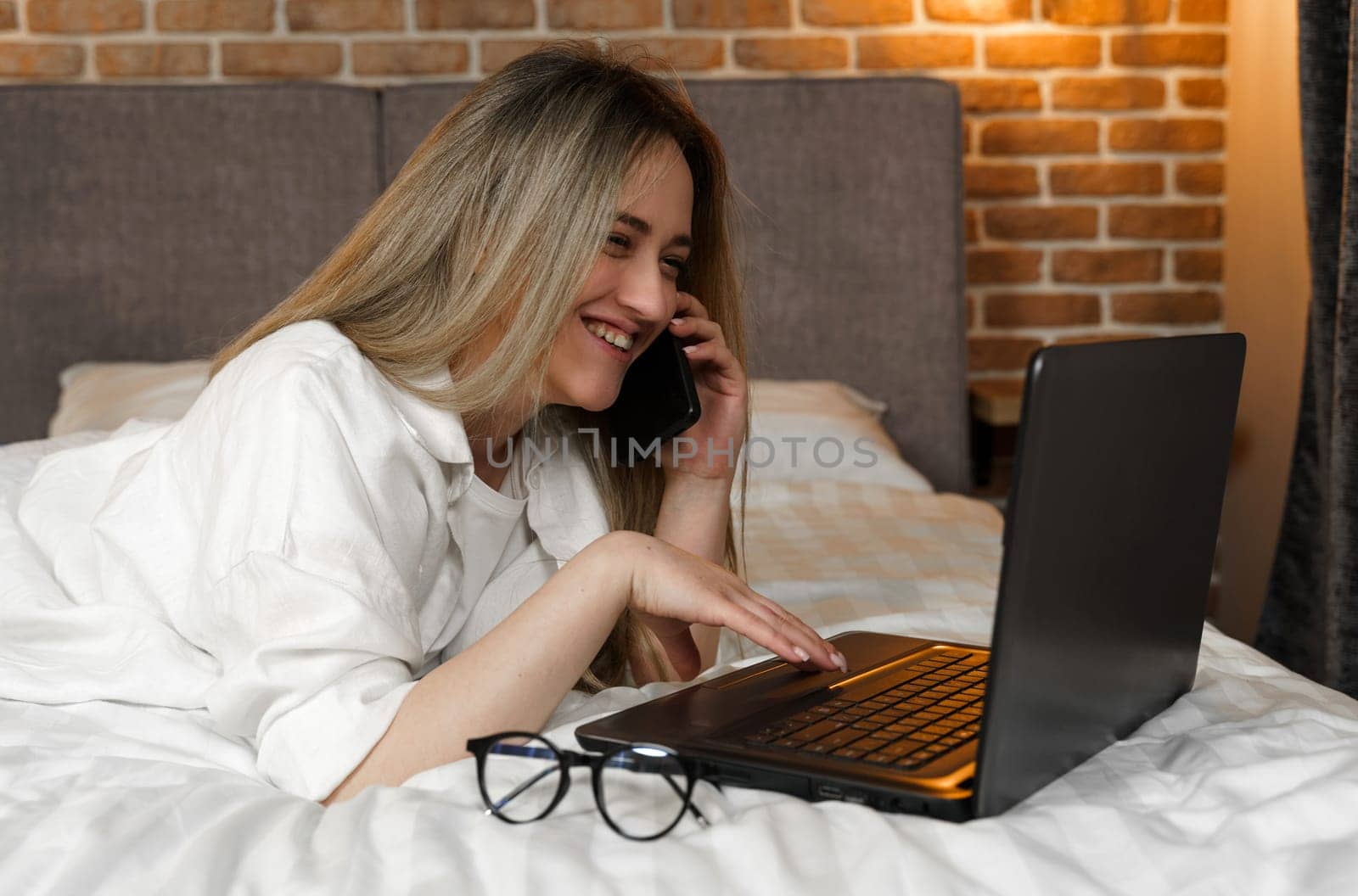 A young beautiful woman is lying on the bed, talking on the phone, looking into a laptop.