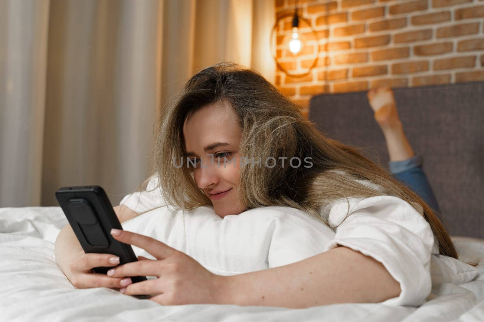A young beautiful woman is lying on the bed, watching content in a smartphone, smiling by Sd28DimoN_1976