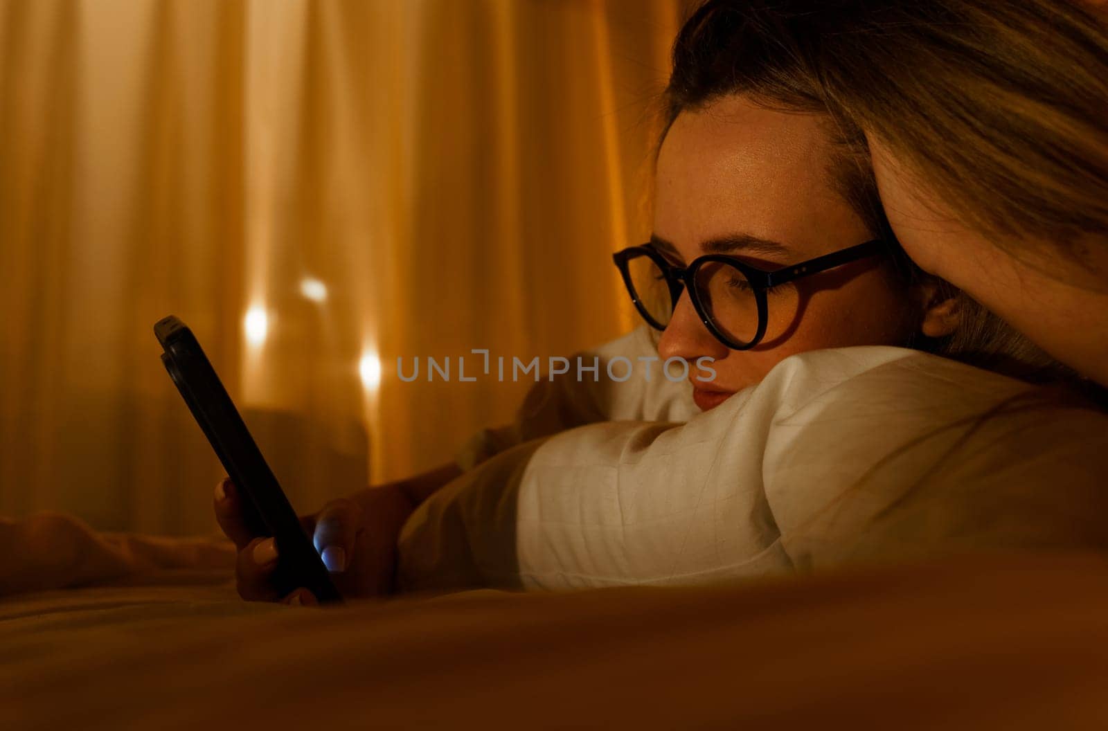 Young woman in glasses with smartphone lying in bed at home at night. Close-up