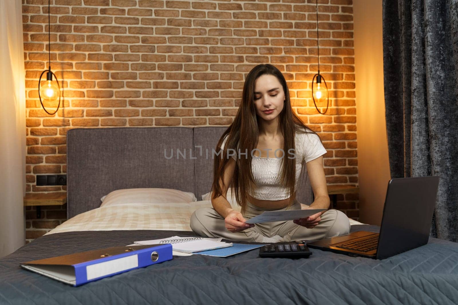 A young woman sits on a bed, looks at reporting documents, counts on a calculator and works on a laptop.