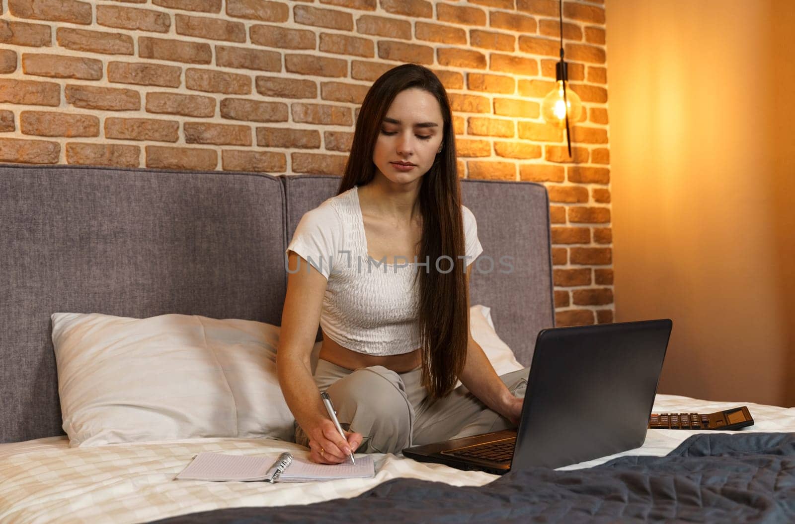 Happy beautiful woman sits on the bed, works on a laptop, makes notes in a notebook by Sd28DimoN_1976