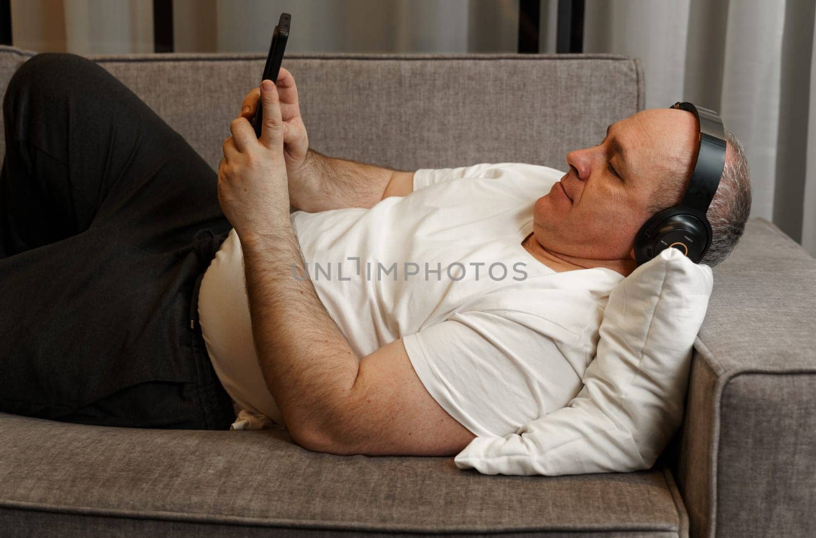 A middle-aged man lies on the couch, listens to music on headphones, looks at the phone.