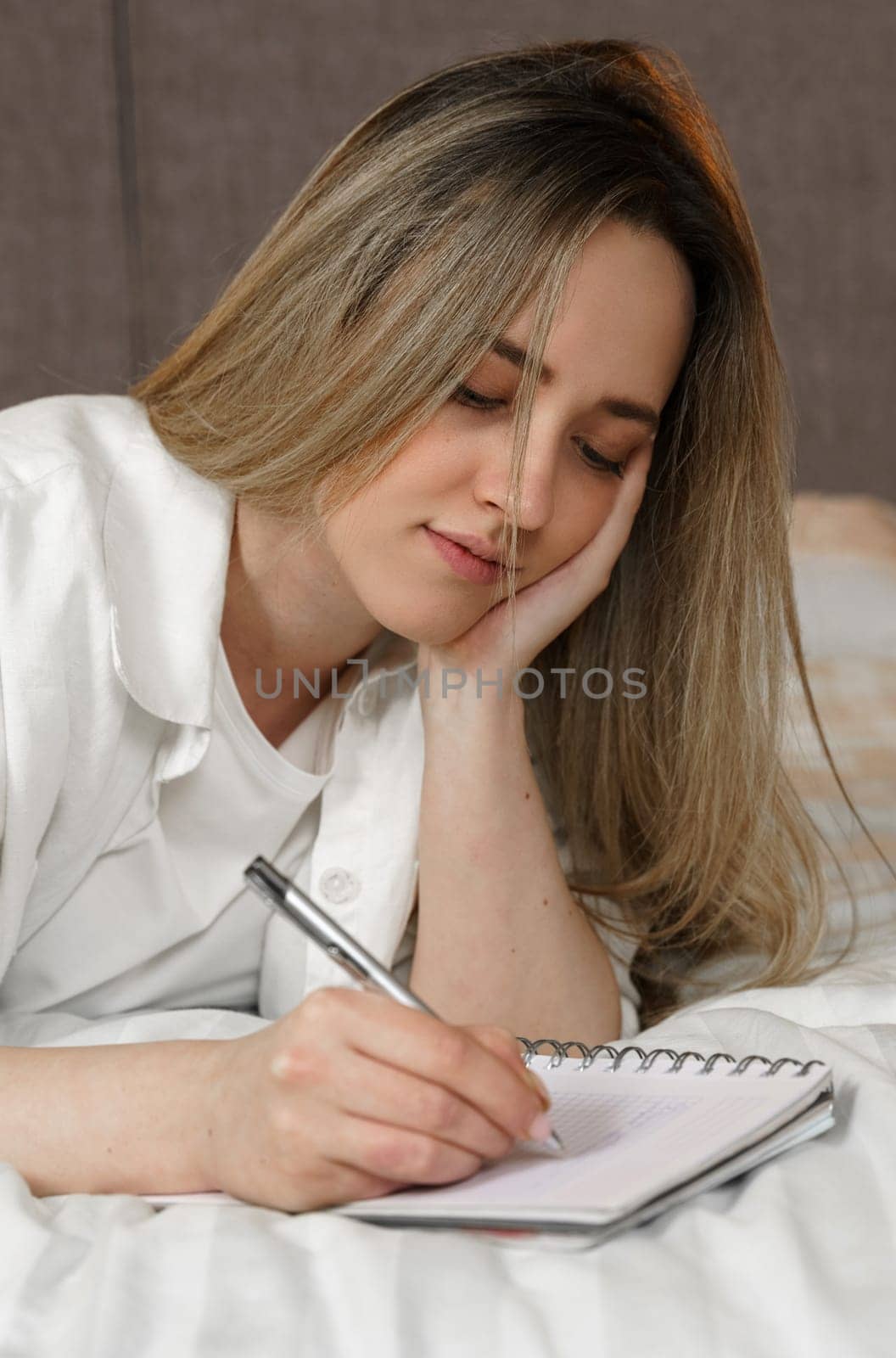 A young beautiful woman lies on the bed and makes notes in a notebook. Vertical frame.