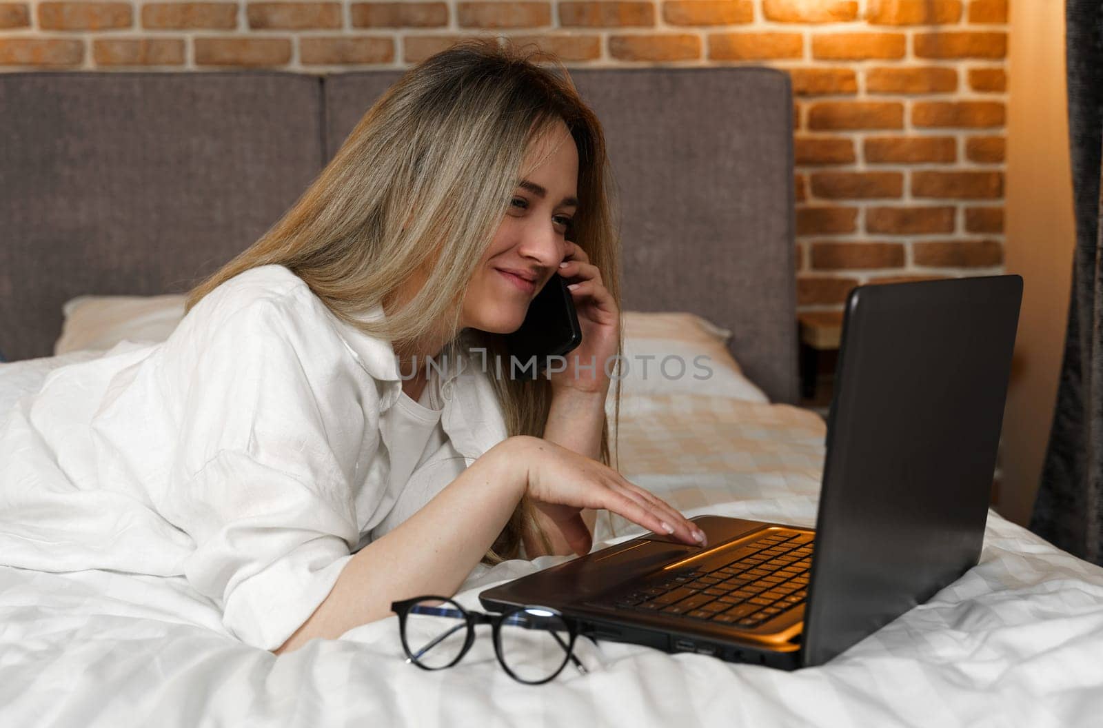 A young beautiful woman is lying on the bed, talking on the phone, looking into a laptop.