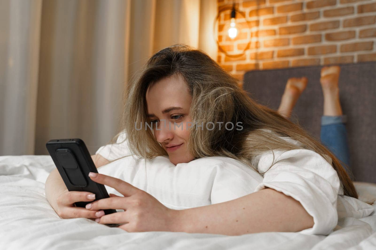 A young beautiful woman is lying on the bed, watching content in a smartphone, smiling by Sd28DimoN_1976