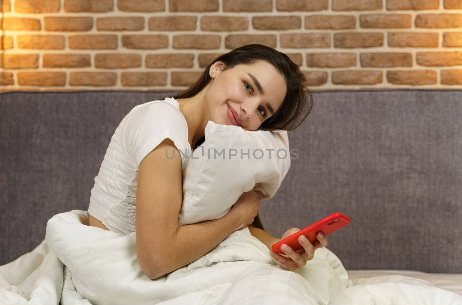 A young beautiful woman sits on a bed hugging a pillow, holding a phone in her hands, looking at the camera.