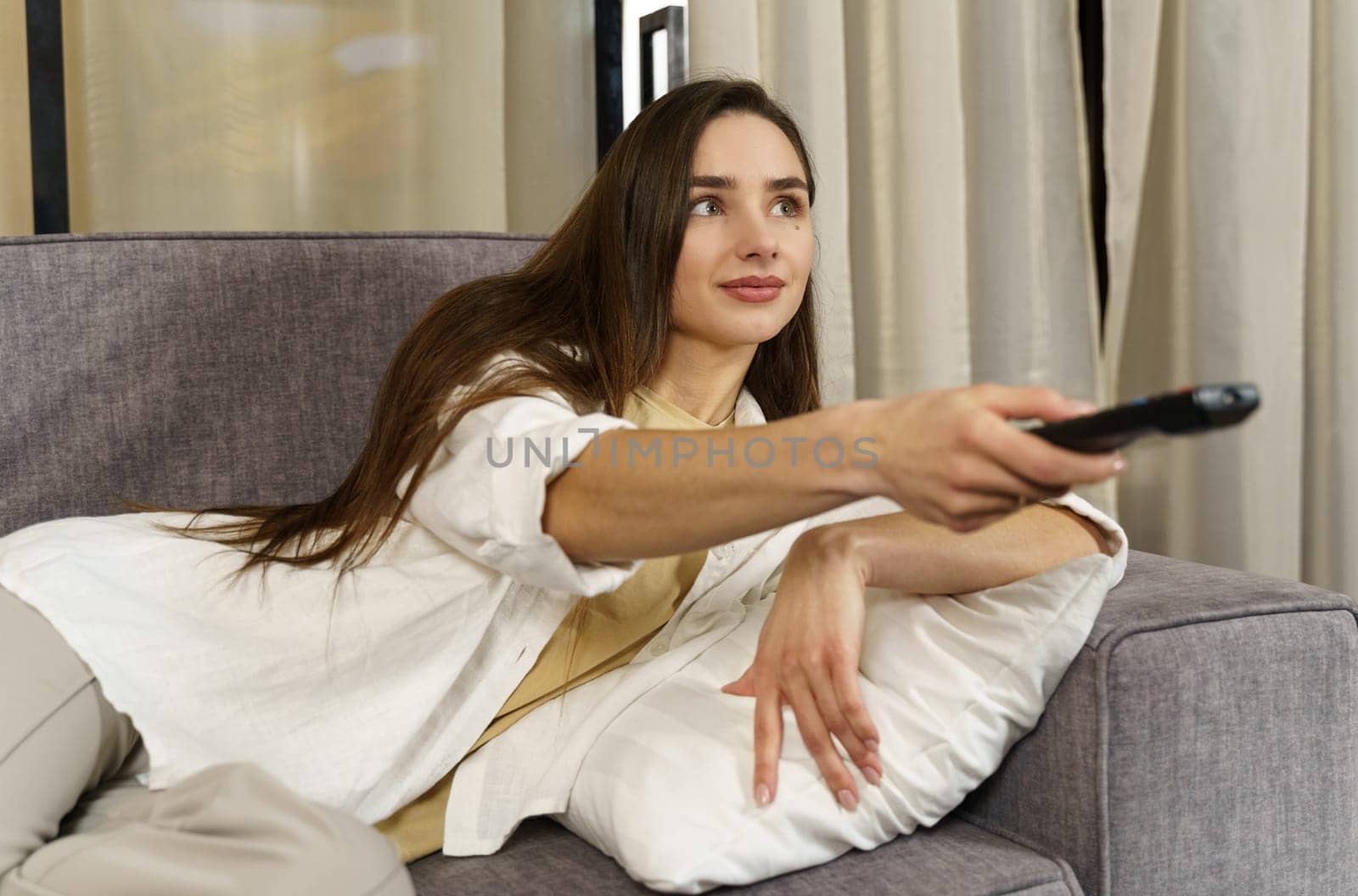 Positive young woman sitting on the sofa indoors at home and watching TV with remote control on the sofa.
