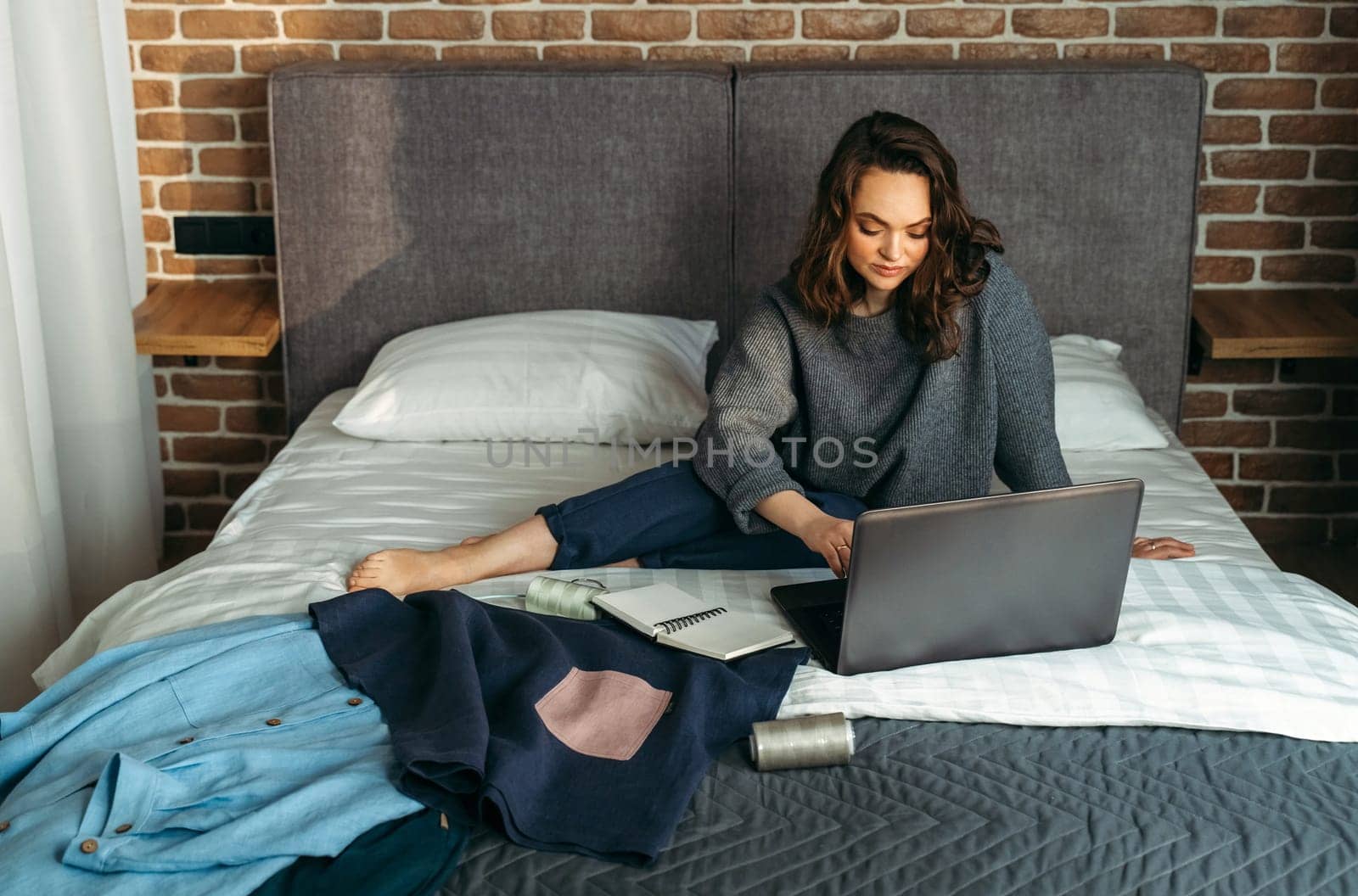 Women's outfits are laid out on the bed, a woman is sitting next to the bed and working at the computer.