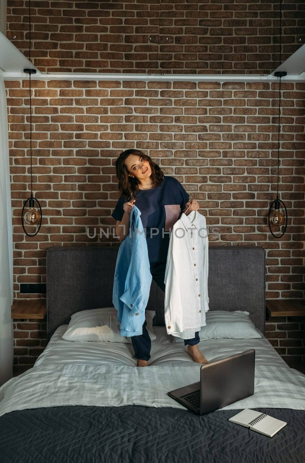 A beautiful woman in designer clothes stands on the bed and holds clothes bought online in her hands. Vertical frame.