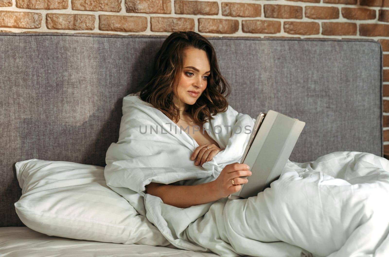 A young woman lies in bed in the morning, watches the news on her tablet.