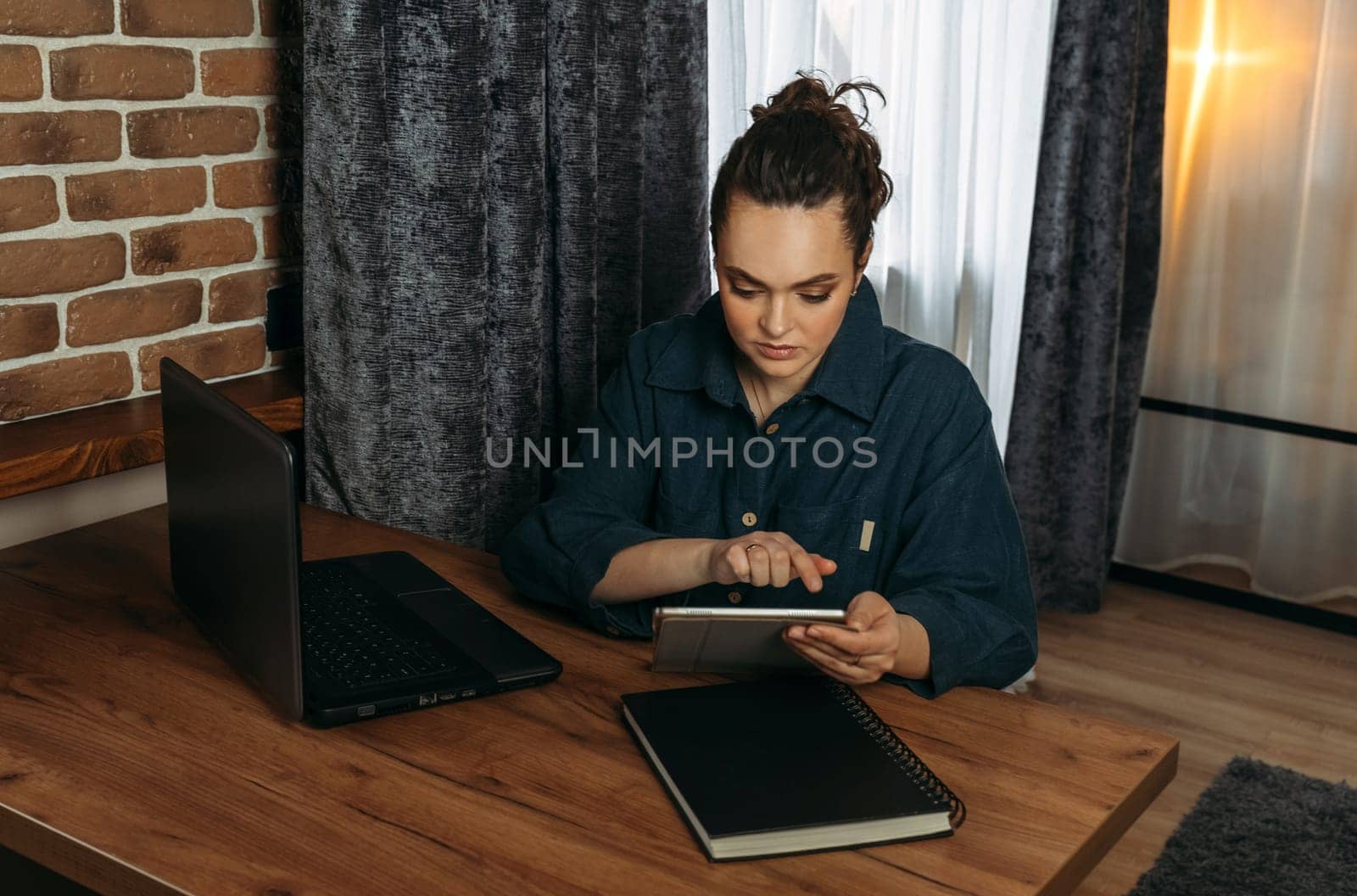 A beautiful woman sits at a table indoors and works remotely on a tablet.