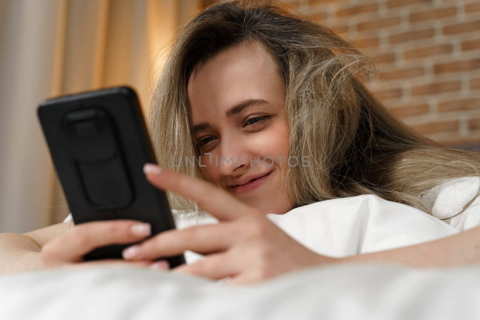 A young beautiful woman is lying on the bed, watching content in a smartphone, smiling by Sd28DimoN_1976