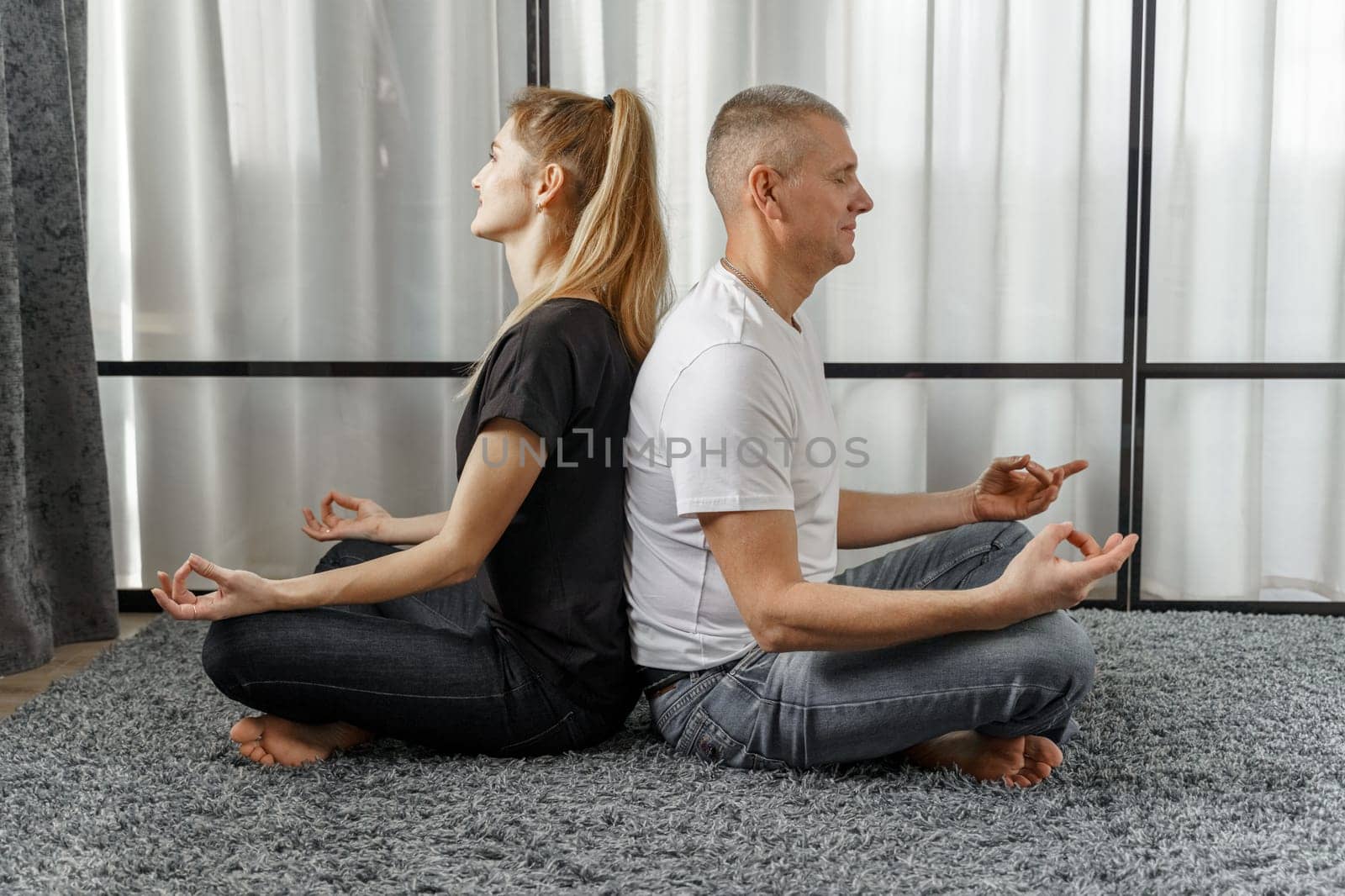 A man and a woman sit back to back, practice yoga and meditate for relaxation and balance of life in a room at home. Sports concept.