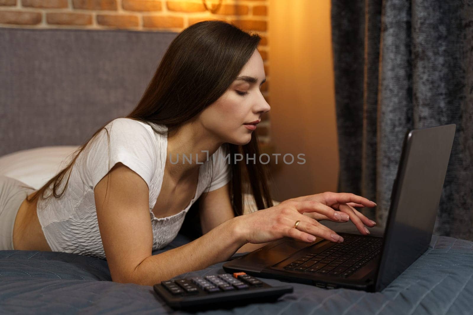 Happy beautiful woman lies on the bed and works on a laptop.