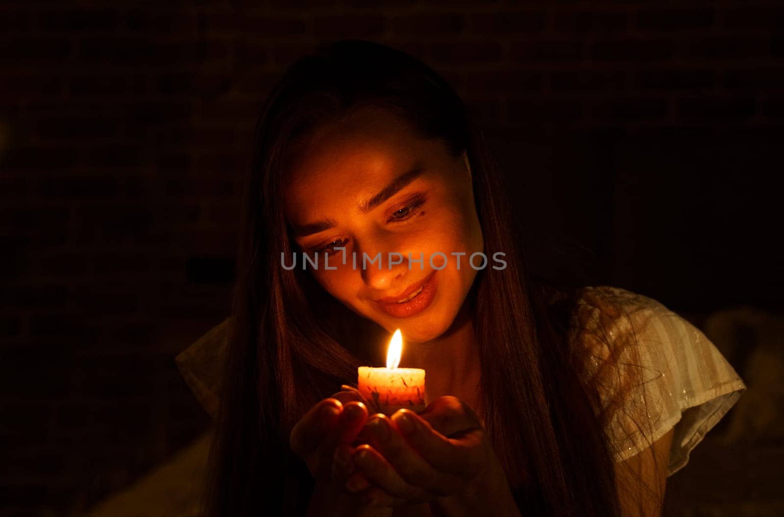 A woman holds a burning candle in her palms on a dark black background.