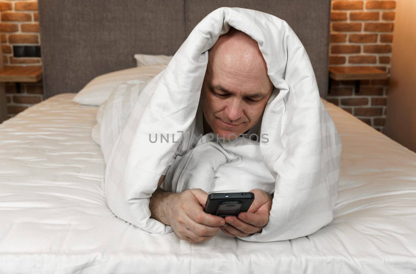 An adult man lies covered with a blanket and browses social networks, videos in a smartphone.