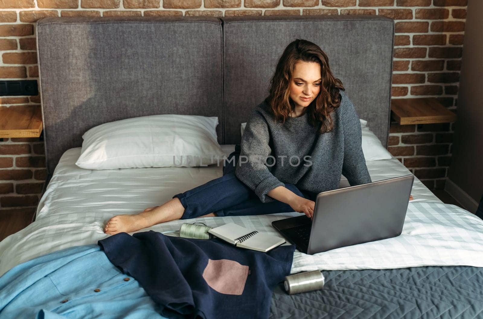 Women's outfits are laid out on the bed, a woman is sitting next to the bed and working at the computer.