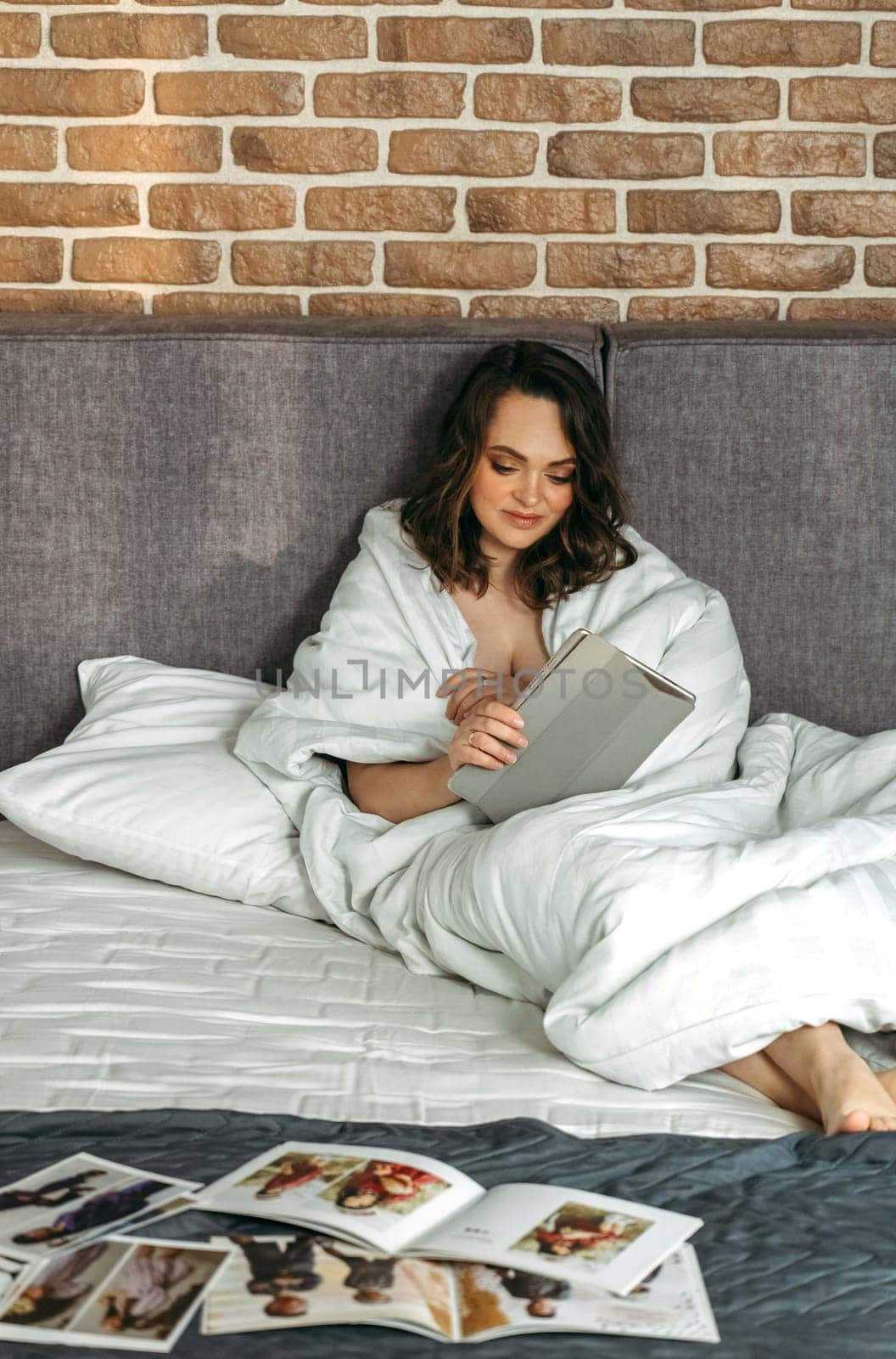 A young woman lies in bed, makes online purchases through a tablet, catalogs with clothes lie in front of her. The directory image is blurry. Vertical frame.