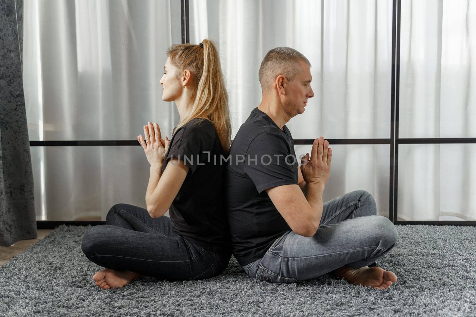 A man and a woman sit back to back, practice yoga and meditate for relaxation and balance of life in a room at home. by Sd28DimoN_1976