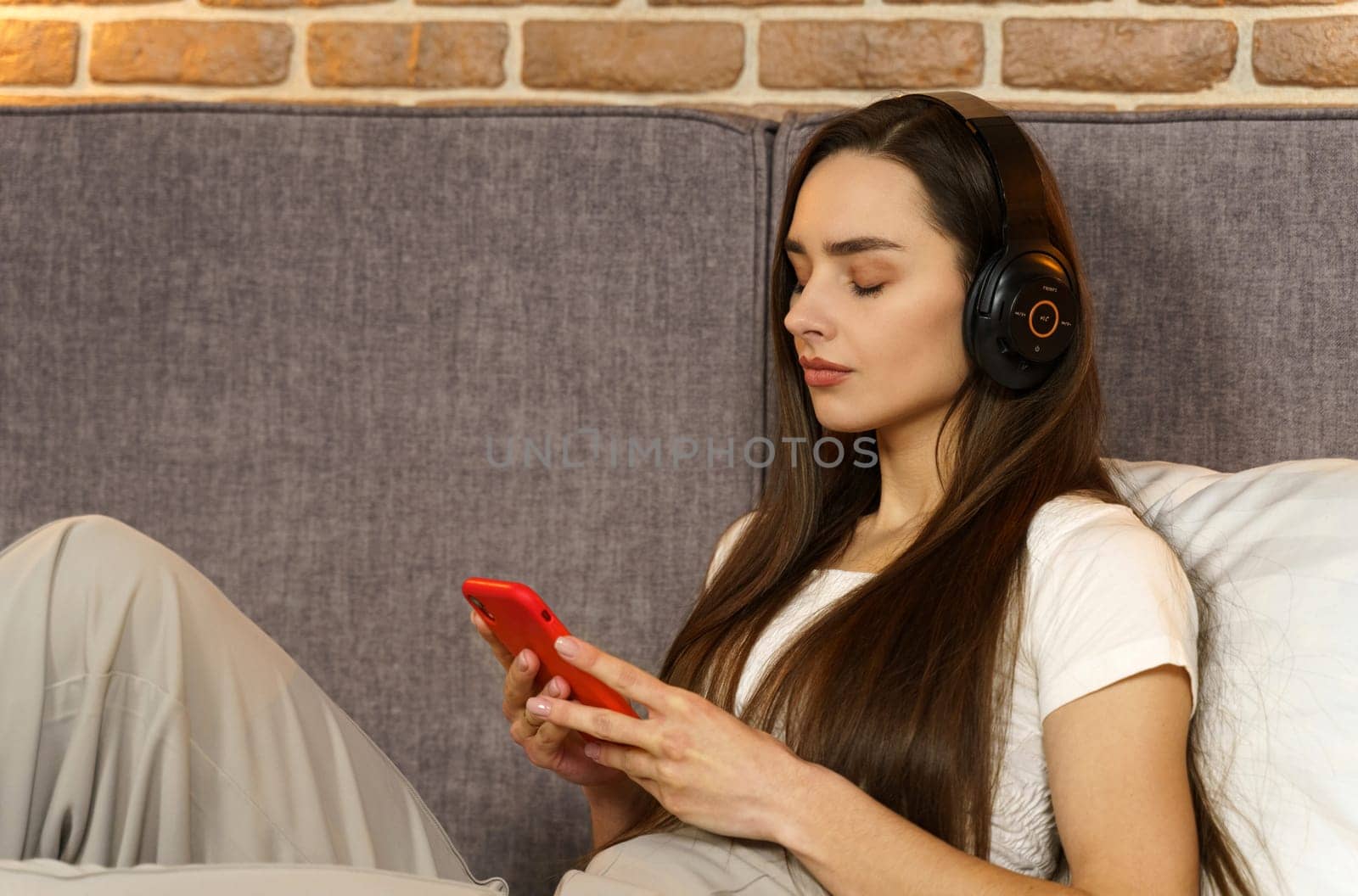 Happy young woman in headphones sits on the sofa and listens to music from a mobile phone, chooses tracks. Side view.