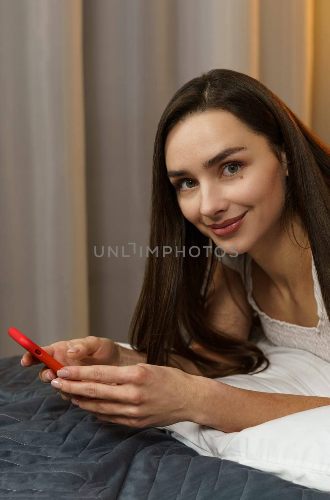 Portrait of a young woman who lies with a smartphone in her hands in the evening on the bed. Looking at the camera. Vertical frame.