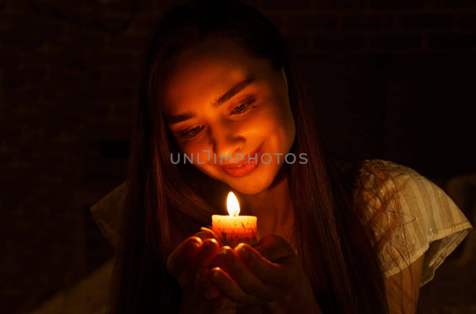 A woman holds a burning candle in her palms on a dark black background.