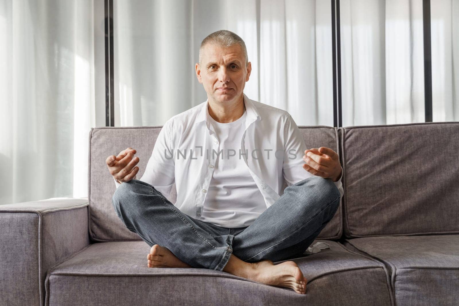 Portrait of a man who sits on a sofa in a room in a lotus position. The man is doing yoga.