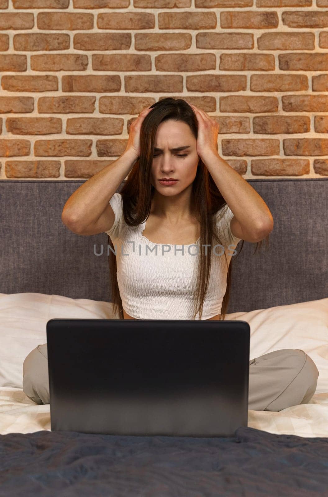 Young business woman with a headache, uses a laptop, works late at night on the bed. Vertical frame.