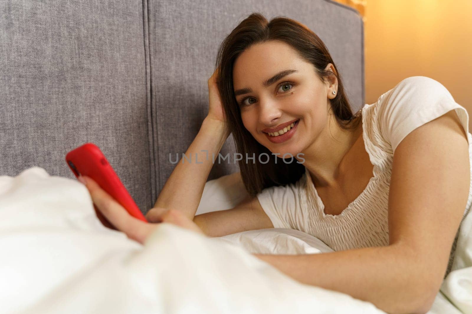 A beautiful young girl lies in bed and enjoys communication, holds a smartphone in her hands, looks at the camera.