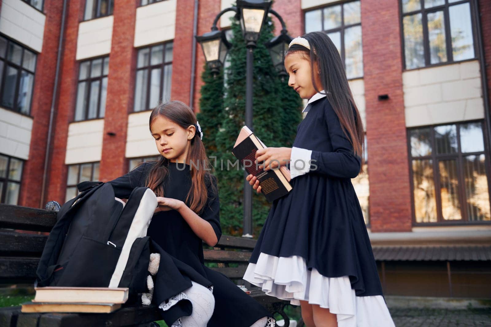 Sitting together. Two schoolgirls is outside near school building.