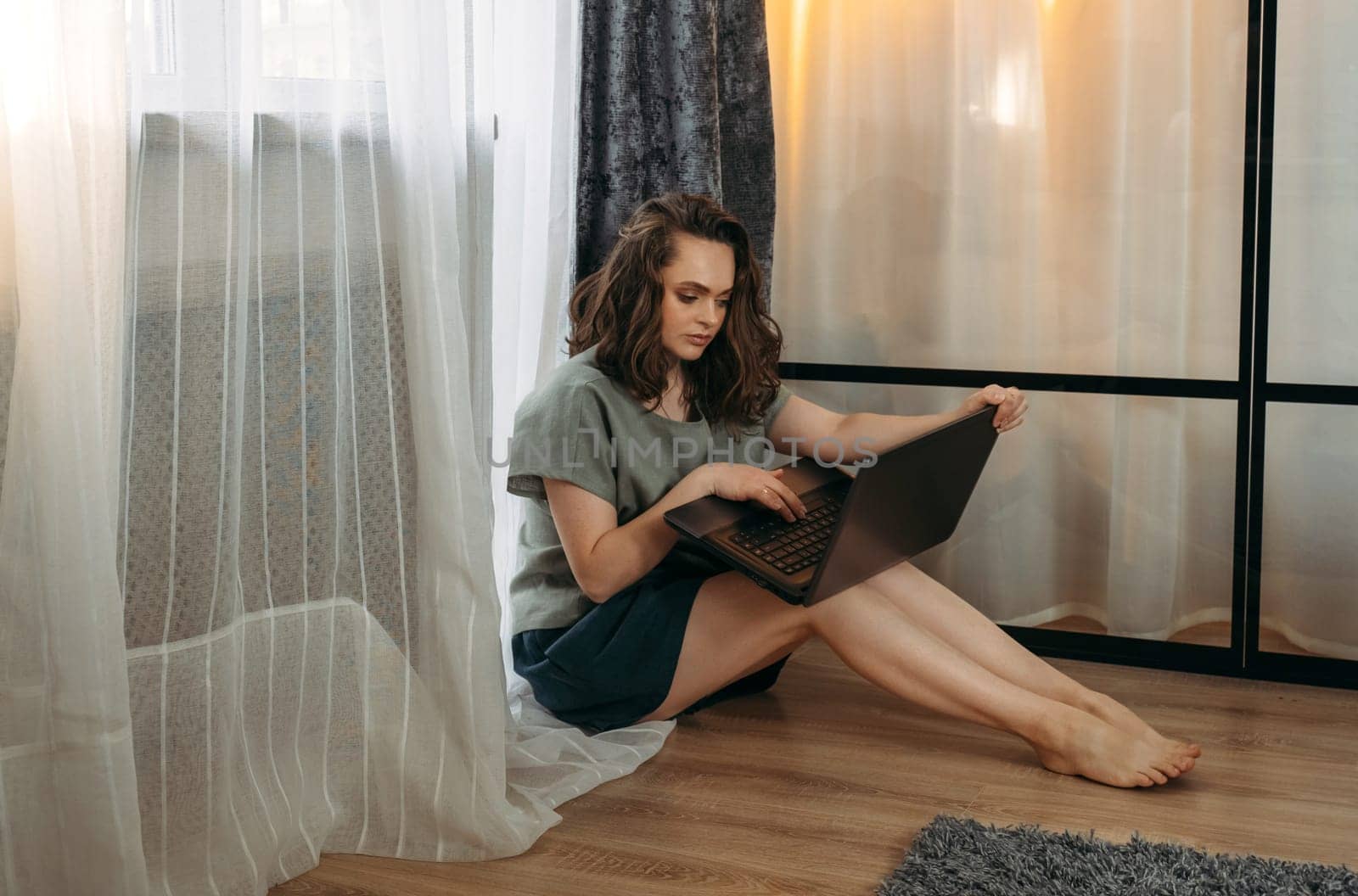 A beautiful woman is sitting on the floor near the window with a laptop, working or studying.