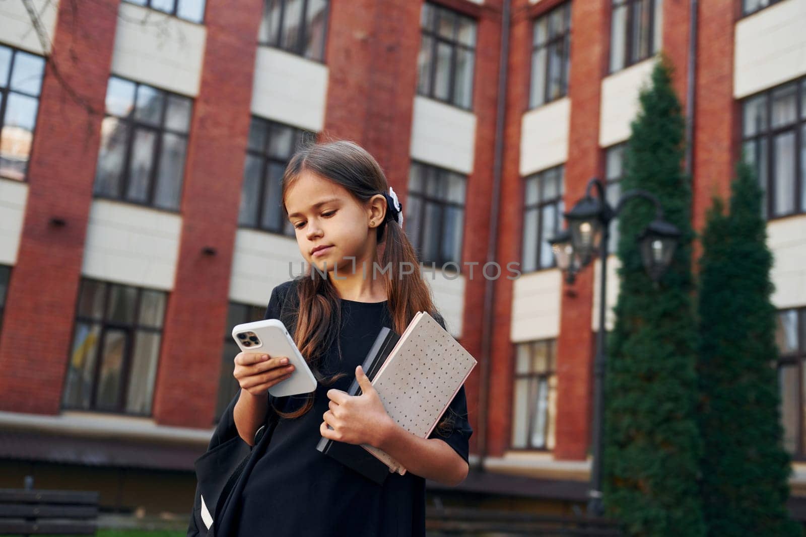Holding phone in hands. Schoolgirl is outside near school building.