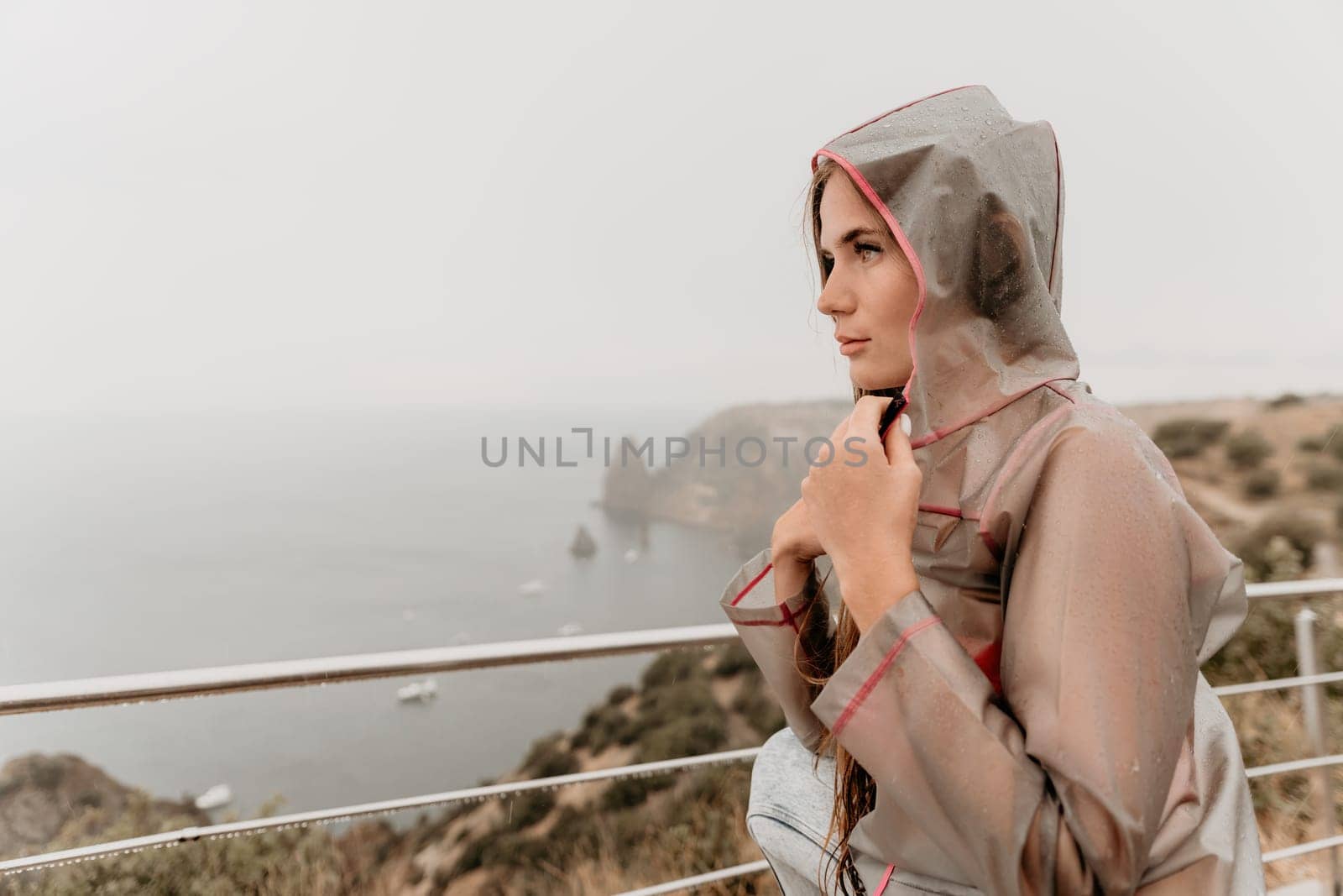 Woman rain park. Happy woman portrait wearing a raincoat with transparent umbrella outdoors on rainy day in park near sea. Girl on the nature on rainy overcast day