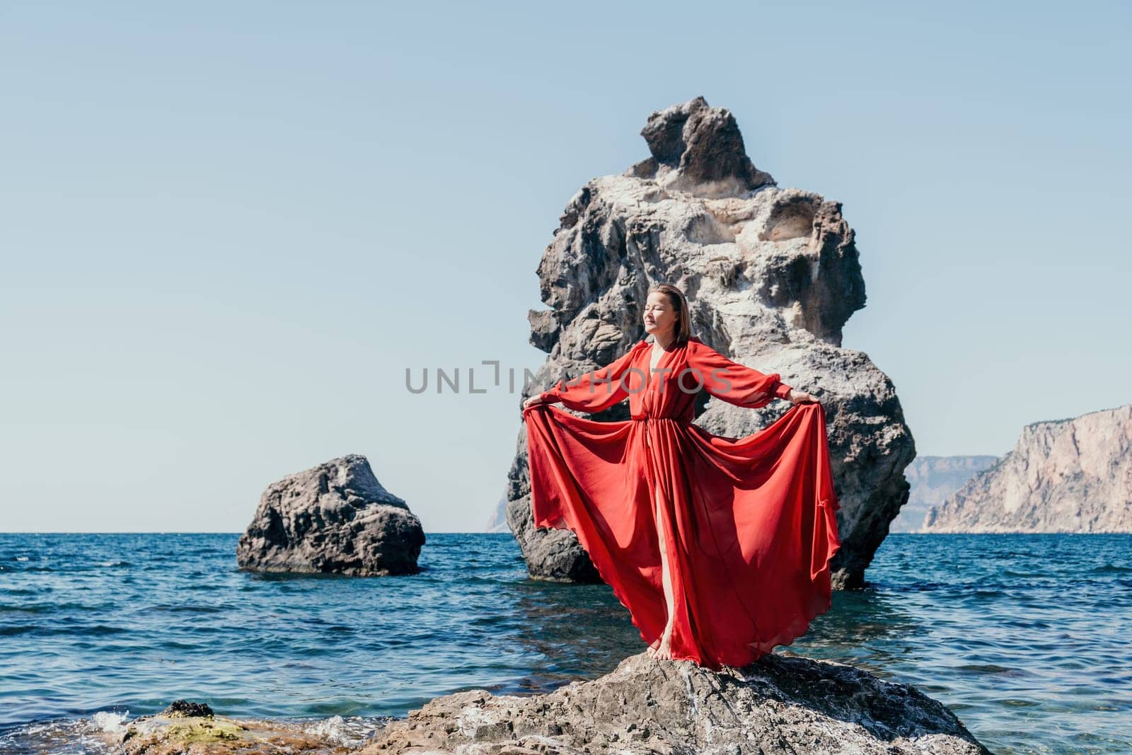 Woman travel sea. Young Happy woman in a long red dress posing on a beach near the sea on background of volcanic rocks, like in Iceland, sharing travel adventure journey