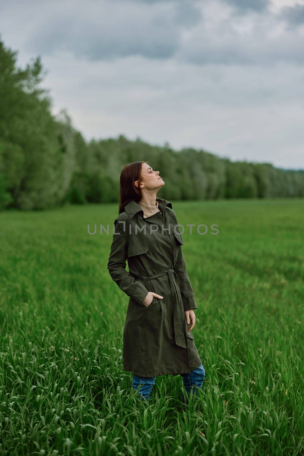 a beautiful woman in a dark coat stands in a green field in the spring in rainy weather. High quality photo