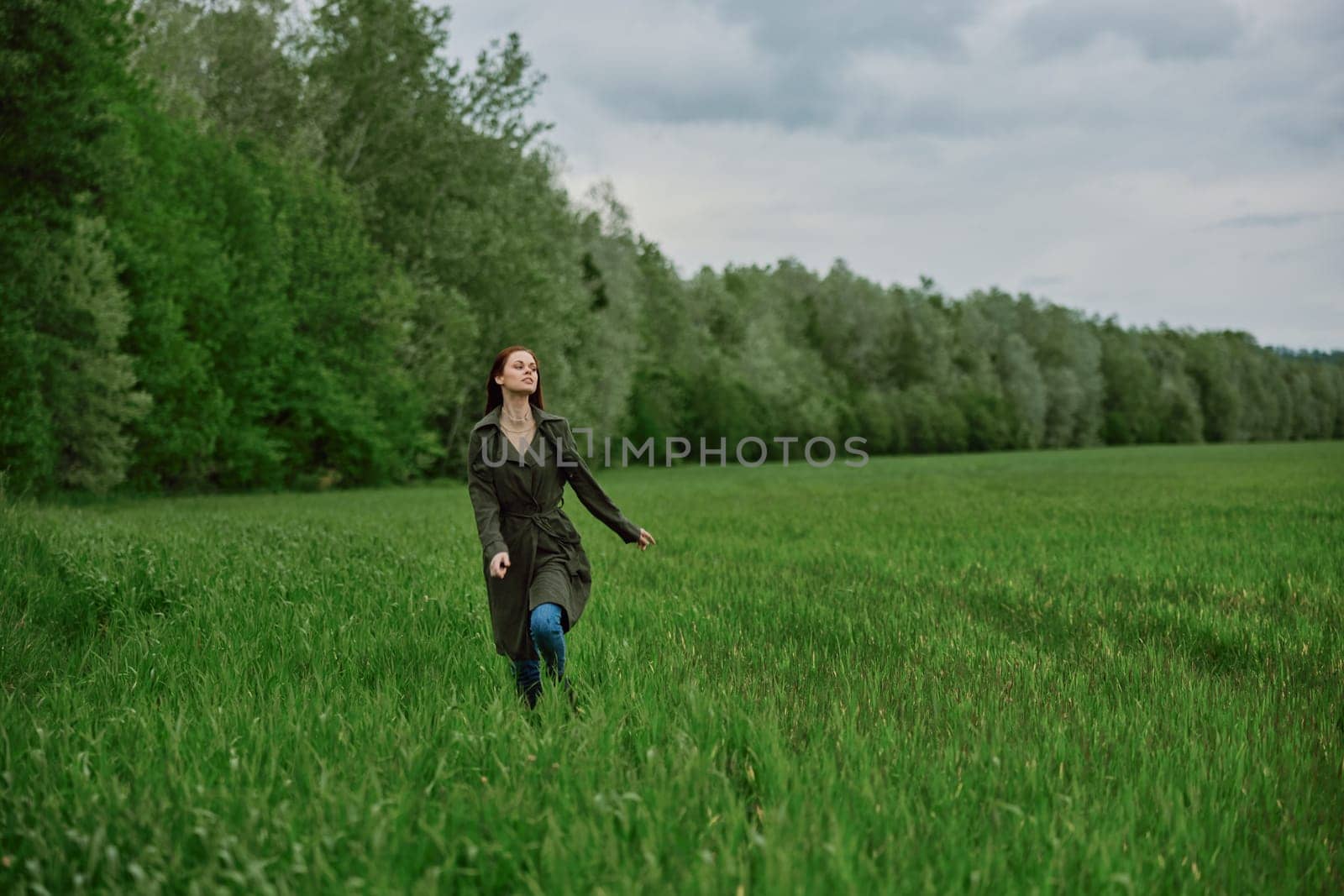 a beautiful woman in a long raincoat runs across a field in high grass in spring in cloudy weather. High quality photo